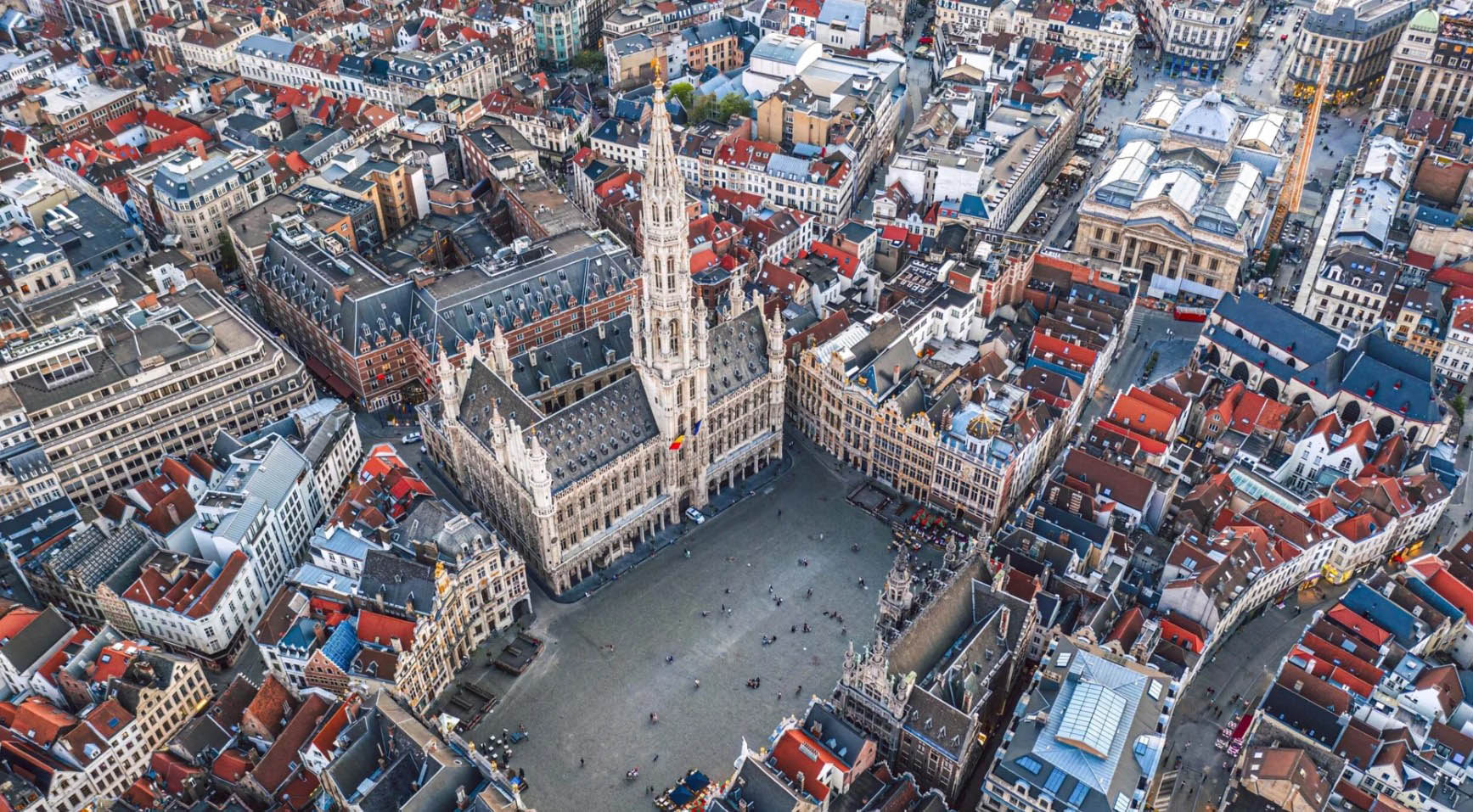 Aerial view of the Grand Place in Brussels, Belgium, surrounded by historic guild houses and ornate civic buildings. The Gothic-style Brussels Town Hall, with its distinctive central spire, dominates the square at the center of the image. Dense urban blocks with red-tiled rooftops and narrow streets extend in all directions, highlighting the city’s historic architecture and compact urban layout. The photograph provides a detailed overview of one of Brussels’ most iconic landmarks.