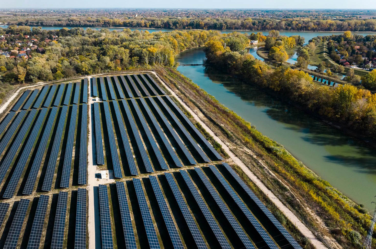 Aerial view of a solar power plant in Hungary, featuring long rows of photovoltaic panels arranged in a grid pattern beside a winding river. Surrounded by trees, waterways, and rural settlements, the installation is integrated into a natural landscape. The image highlights renewable energy generation, with the contrast between the solar farm, green vegetation, and water channels illustrating the coexistence of clean energy infrastructure and the environment.