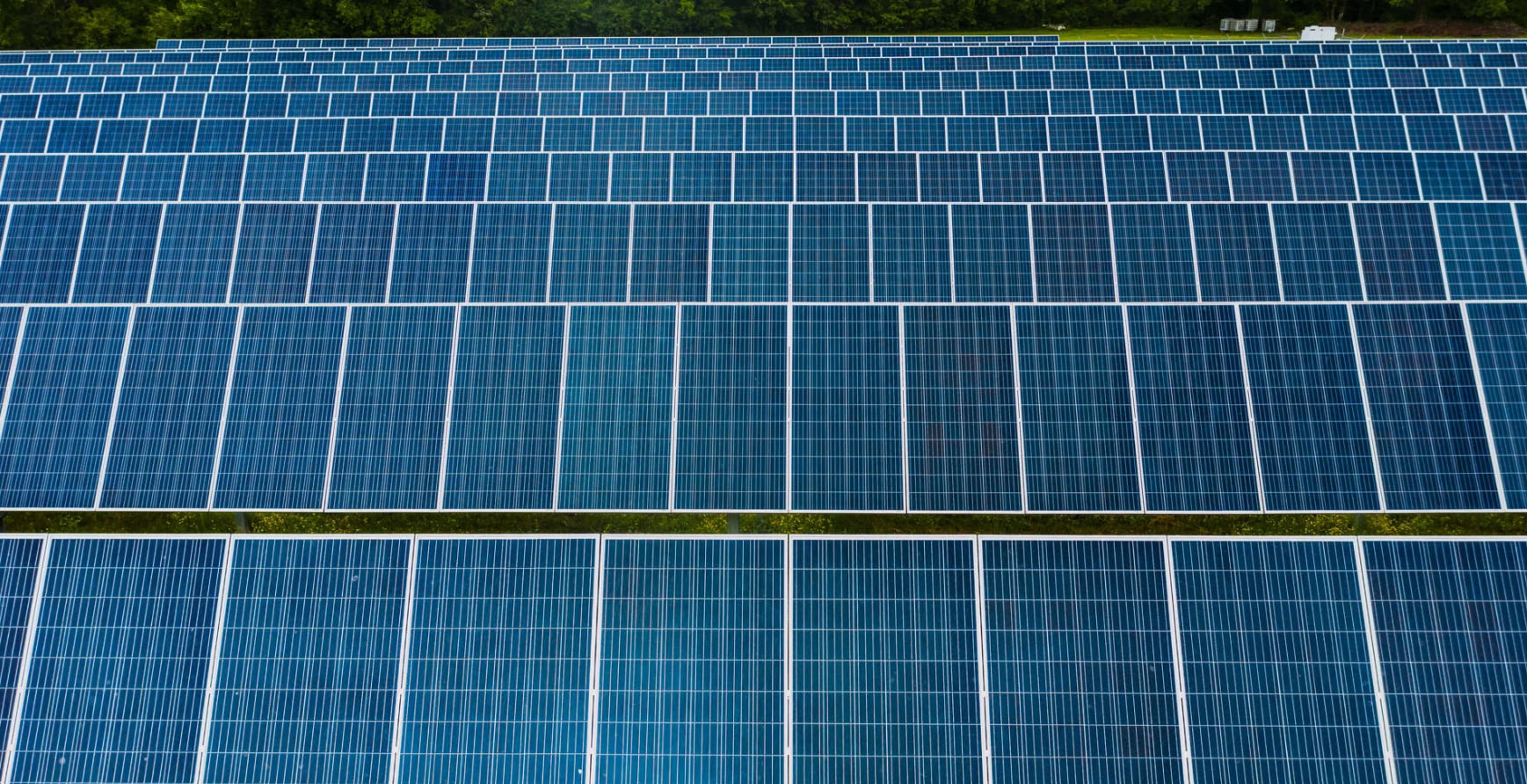 Close-up aerial view of a solar power plant covered with densely arranged photovoltaic panels. The blue solar modules are organized in long, uniform rows that fill almost the entire frame, creating a geometric pattern of repeating rectangles. Narrow green strips of vegetation separate the rows, while a tree line is visible along the upper edge of the image. The photograph emphasizes the scale, efficiency, and structured layout of renewable energy infrastructure.