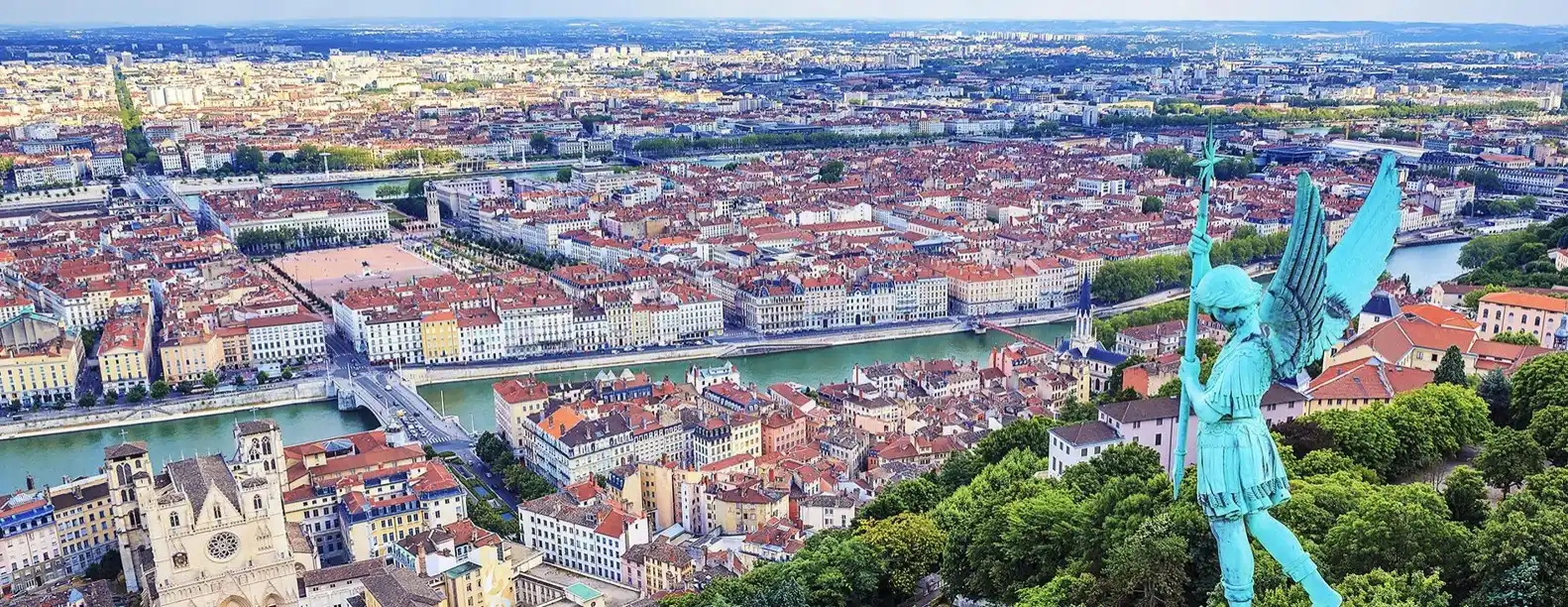 Panoramic aerial view of Lyon, France, showing the city’s dense urban landscape along the river. In the foreground, the rooftop statue near the Basilica of Notre-Dame de Fourvière overlooks historic buildings, bridges, and tree-lined riverbanks. The city extends across the horizon with a mix of historic architecture and modern development, highlighting Lyon’s distinctive geography at the confluence of two rivers.