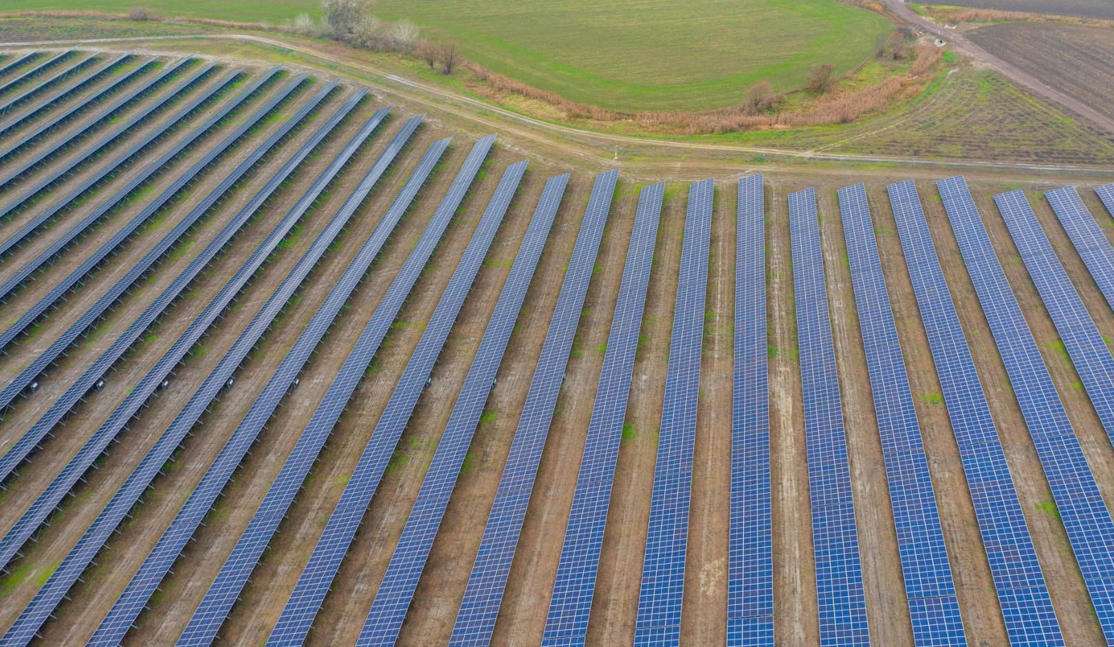 Aerial view of a large solar power plant featuring long, parallel rows of photovoltaic panels extending across open farmland. The solar arrays are arranged in a highly structured grid pattern with maintenance tracks running between them. Surrounding agricultural fields and grassy areas frame the installation, emphasizing the scale of the renewable energy facility and its integration into a rural landscape.